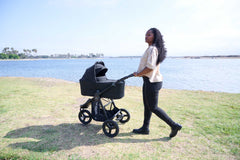 Mother looking into the distance pushing a Bumbleride Indie All Terrain Stroller with Bassinet in Black attached on grass with bay in background. Global